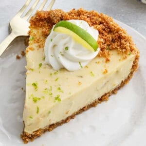 A slice of Key Lime Pie with a graham cracker crust, topped with a swirl of whipped cream and a small slice of lime, served on a white plate with a gold fork.