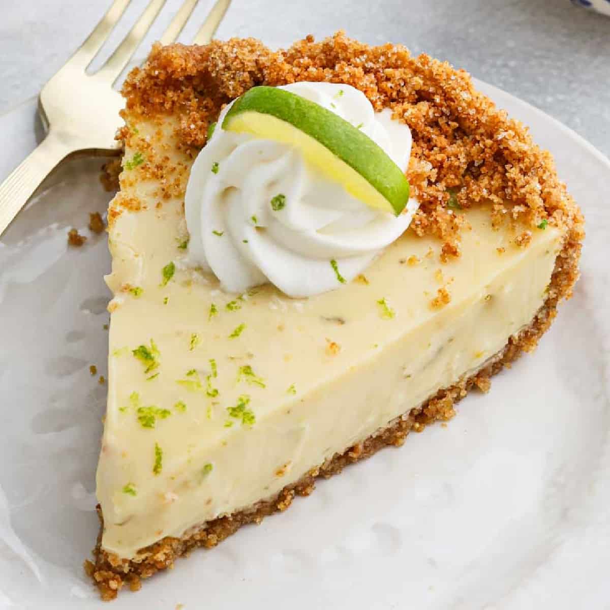 A slice of Key Lime Pie with a graham cracker crust, topped with a swirl of whipped cream and a small slice of lime, served on a white plate with a gold fork.