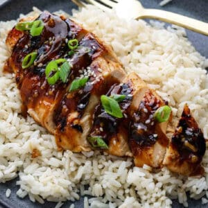 Sliced Korean BBQ Chicken breast topped with dark sauce, sesame seeds, and chopped green onions, served on a bed of white rice on a plate with a fork.