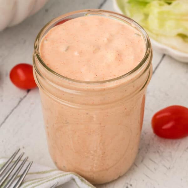 A glass jar filled with creamy, pinkish-orange Thousand Island Dressing sits on a white wooden surface, surrounded by grape tomatoes, lettuce, and a fork with a striped napkin.