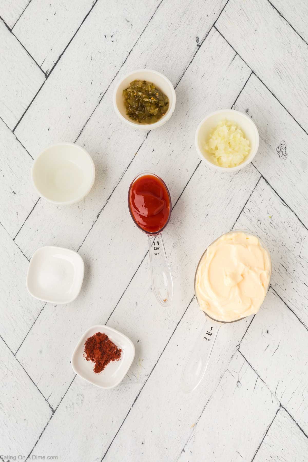 Seven small bowls and measuring spoons on a white wooden surface hold the classic ingredients for a creamy dressing like Thousand Island Dressing—mayonnaise, ketchup, chopped onions, relish, paprika, vinegar, and salt—arranged in a loose circle.