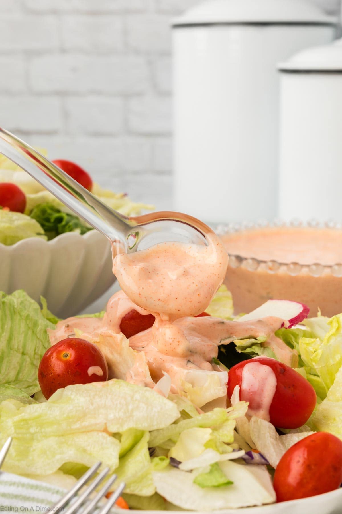 A glass spoon drizzles creamy pink Thousand Island Dressing over a fresh lettuce salad with cherry tomatoes and radish slices, with a bowl of dressing and white canisters in the background.