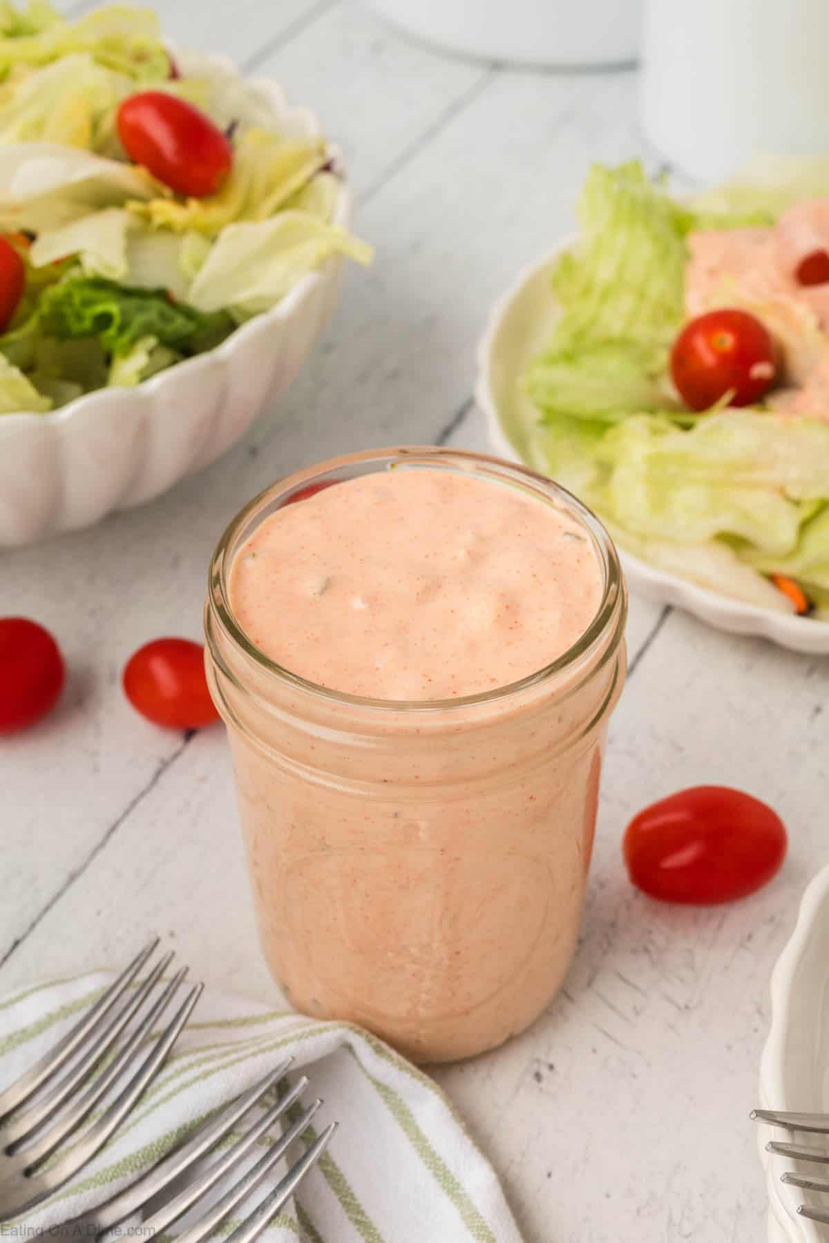 A glass jar of creamy pink Thousand Island Dressing sits on a white wood table, surrounded by fresh salads with lettuce and cherry tomatoes, loose tomatoes, and silver forks—an inviting condiment for any meal.