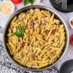 A skillet filled with creamy Chicken Pesto Pasta—penne, chunks of chicken, sun-dried tomatoes, and herbs—garnished with fresh basil. Surrounded by a bowl of grated cheese, tomatoes, plates, and utensils.