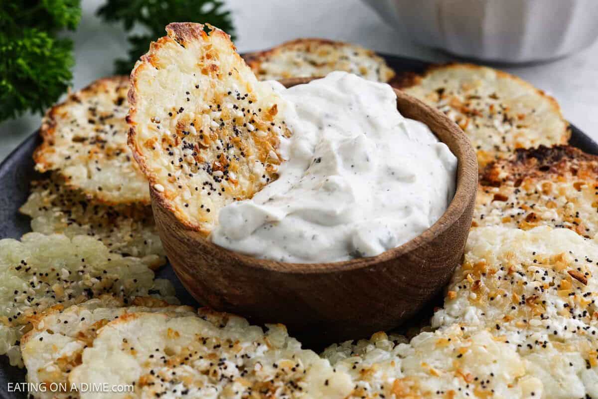 A plate of crispy Cottage Cheese Chips topped with seasoning surrounds a wooden bowl filled with creamy white dip. One chip is partially dipped into the bowl.