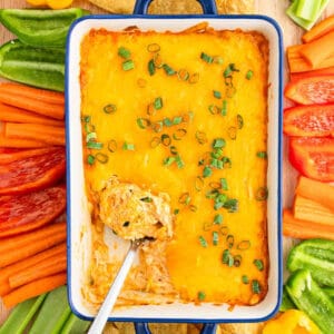 A casserole dish filled with Cottage Cheese Buffalo Chicken Dip topped with chopped green onions, surrounded by sliced carrots, red bell peppers, green bell peppers, and tortilla chips. A spoon scoops dip from the corner.