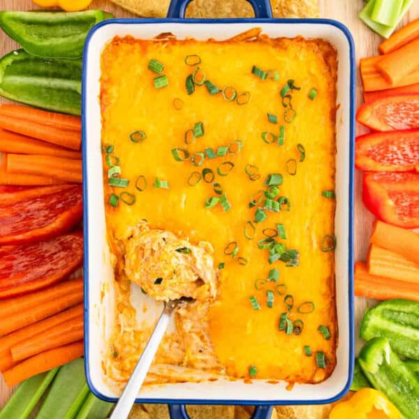 A casserole dish filled with Cottage Cheese Buffalo Chicken Dip topped with chopped green onions, surrounded by sliced carrots, red bell peppers, green bell peppers, and tortilla chips. A spoon scoops dip from the corner.