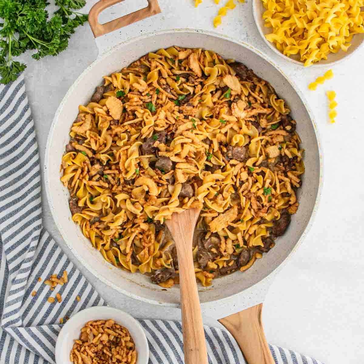A skillet filled with French Onion Beef and Noodles, garnished with herbs, sits on a striped towel. A wooden spoon rests inside, while uncooked noodles, fried onions, and fresh parsley are arranged nearby.