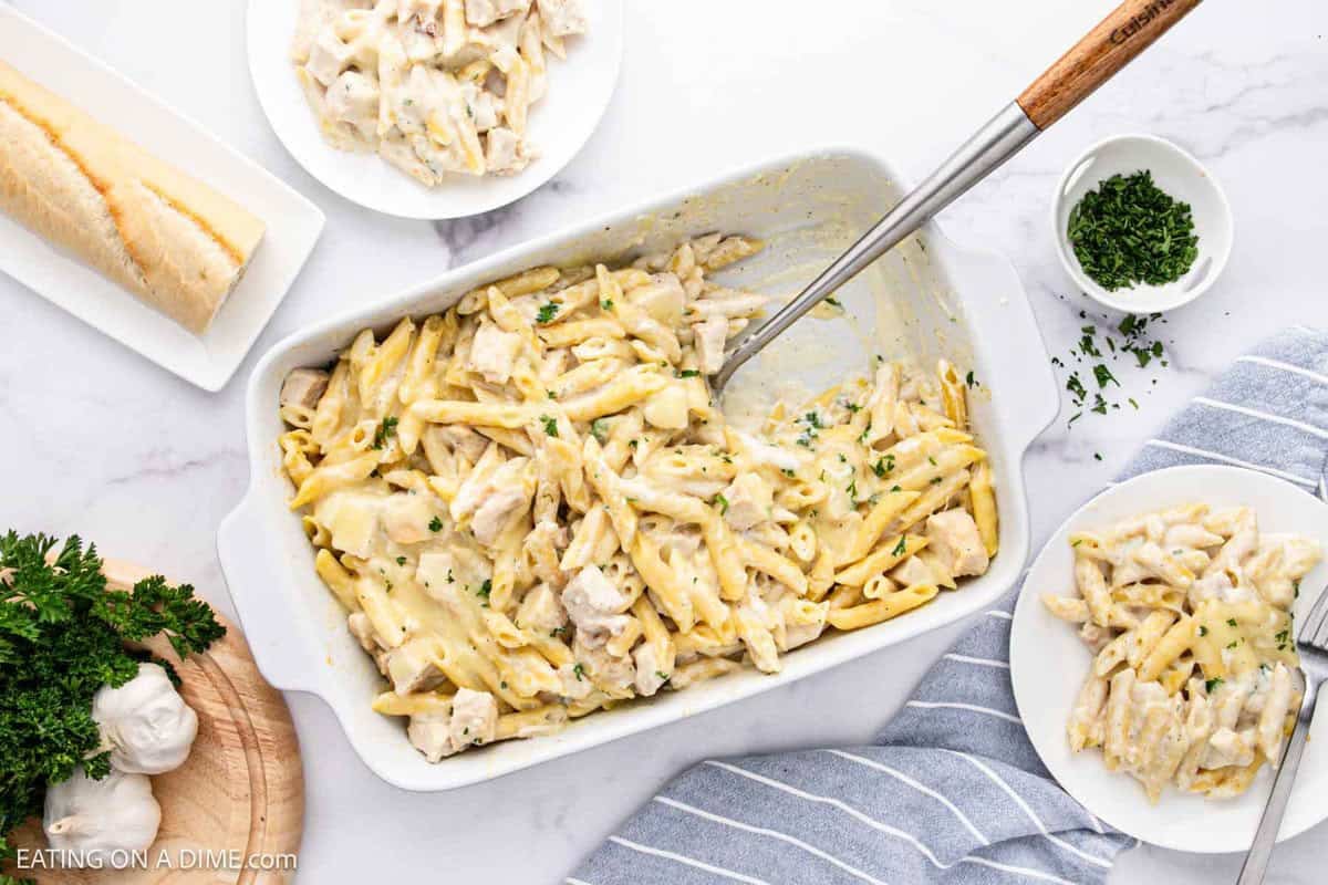 A Chicken Alfredo Bake filled with creamy penne pasta sits on a table, surrounded by two small plates of pasta, a loaf of bread, chopped parsley, and a bulb of garlic. A serving spoon rests in the dish.