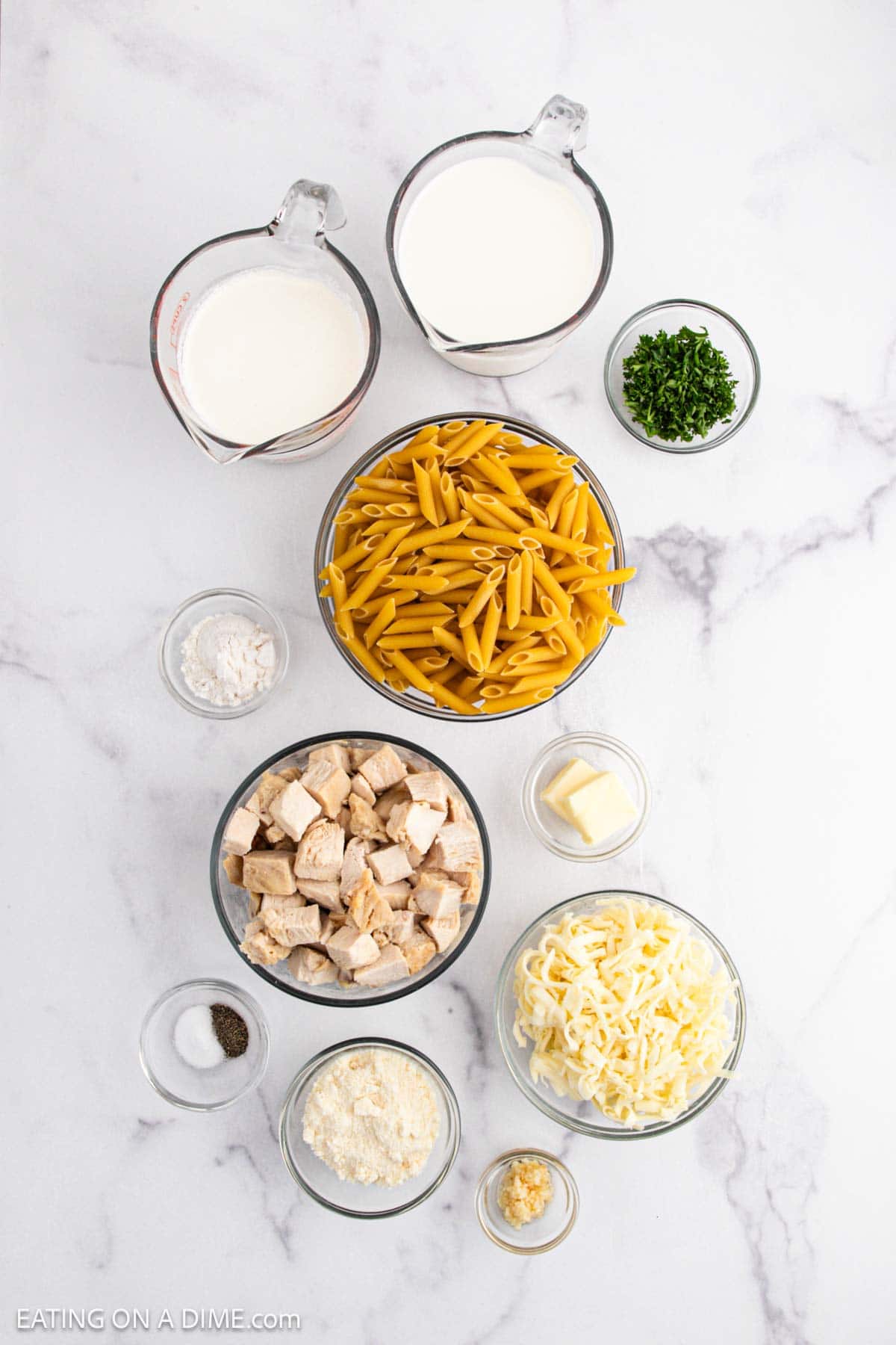 Overhead view of separated Chicken Alfredo ingredients on a white surface: uncooked penne pasta, cubed cooked chicken, shredded cheese, milk, cream, butter, flour, minced garlic, grated parmesan, chopped parsley, and salt and pepper.