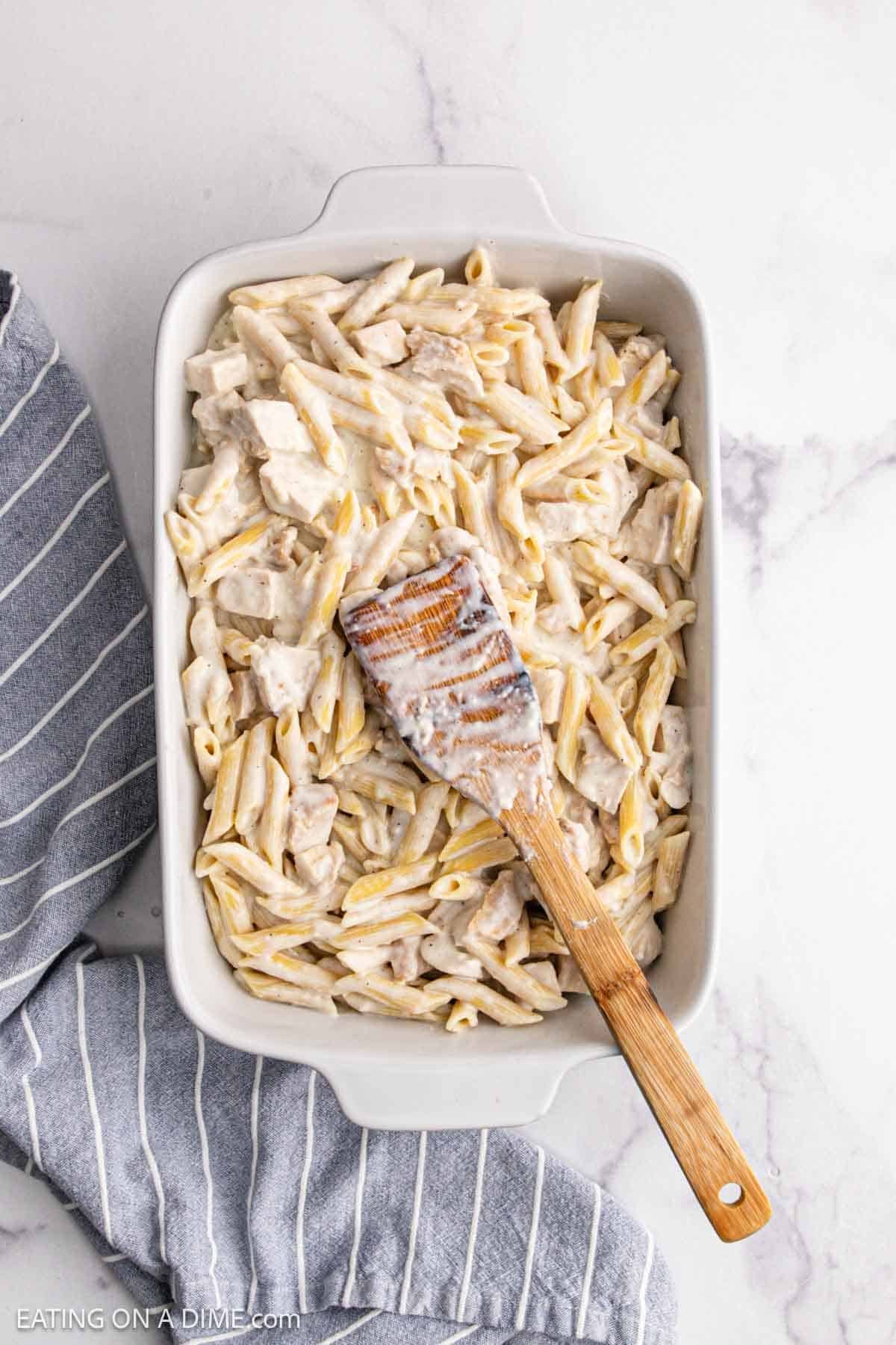 A white baking dish filled with creamy Chicken Alfredo Bake and tender chicken pieces, topped with a wooden spatula coated in sauce. The dish rests on a gray and white striped kitchen towel on a marble countertop.