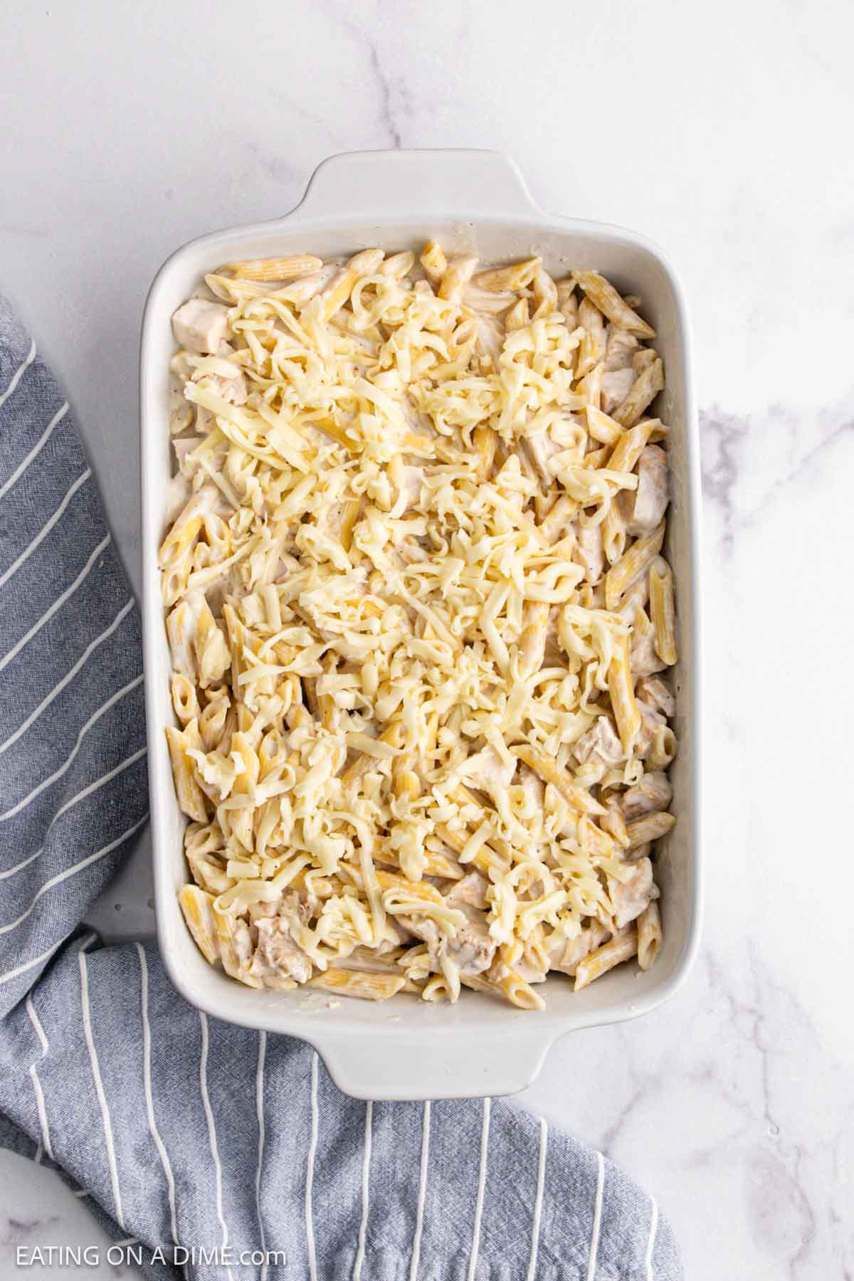 A white baking dish filled with Chicken Alfredo Bake—cooked penne pasta mixed with shredded chicken and cheese—sits on a marble countertop next to a gray and white striped kitchen towel.