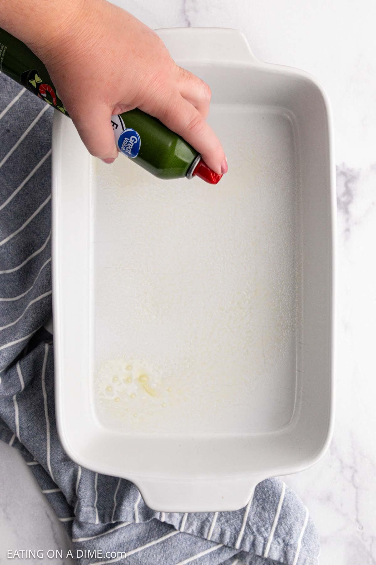 A hand sprays oil from a can onto a large white rectangular baking dish, prepping it for a delicious Chicken Alfredo Bake, all set on a gray and white striped towel atop a marble surface.