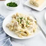 A plate of creamy Chicken Alfredo Bake with chunks of chicken and penne, topped with chopped parsley. A fork rests on the plate, with extra parsley and bread on the side. The setting is bright and casual.
