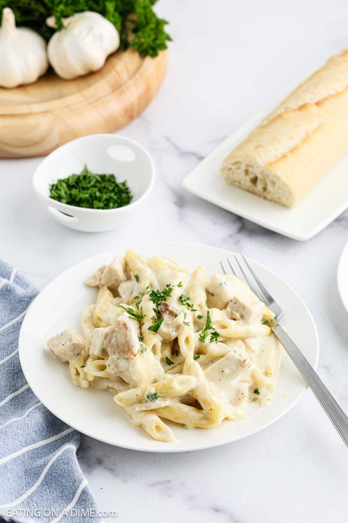 A white plate of creamy Chicken Alfredo Bake penne pasta, garnished with chopped parsley, sits beside a fork. In the background are a bowl of parsley, a loaf of bread, garlic bulbs, and a wooden cutting board.
