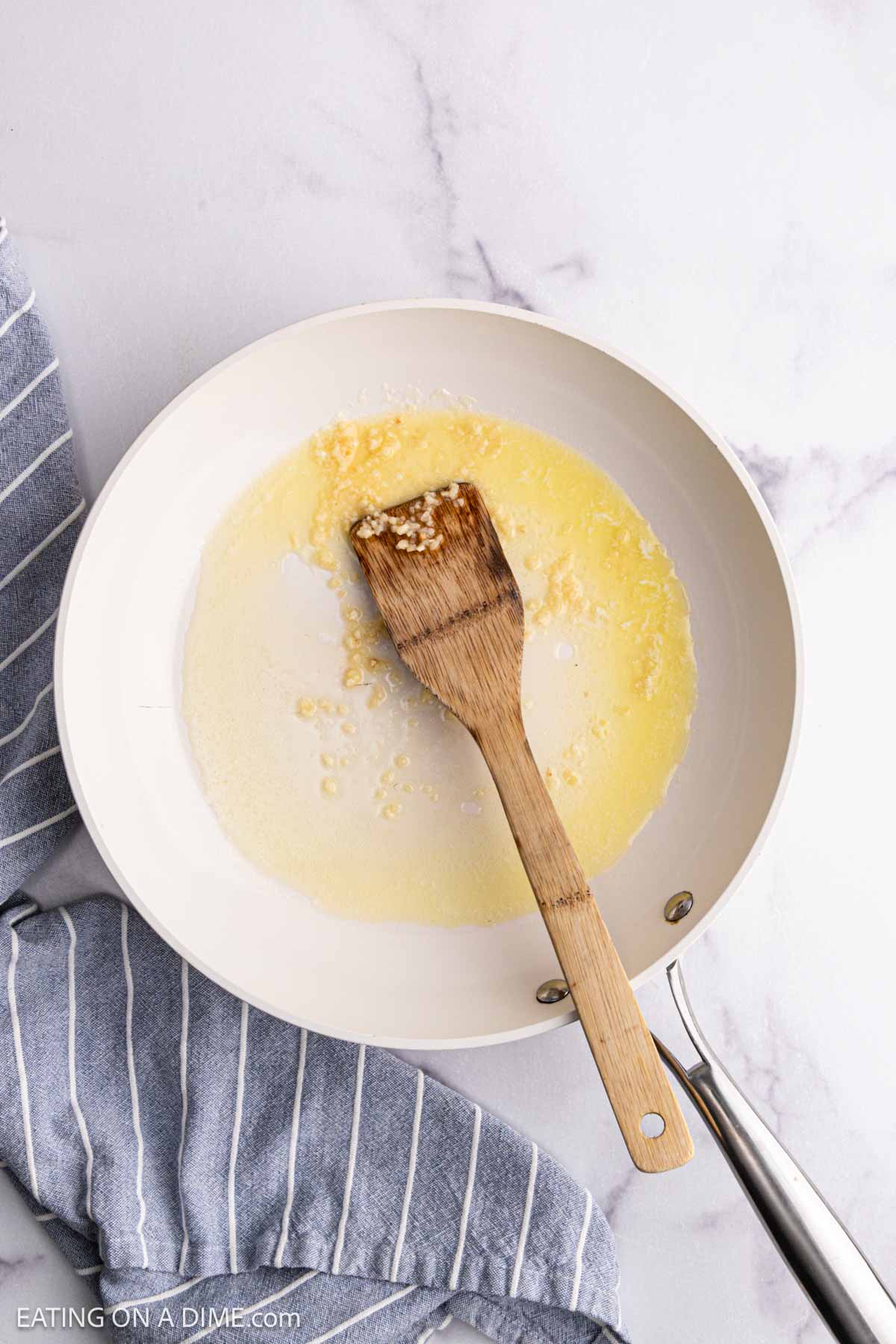 A wooden spatula stirs melted butter and minced garlic in a white skillet on a marble surface, preparing the base for a delicious Chicken Alfredo Bake, with a blue and white striped kitchen towel nearby.