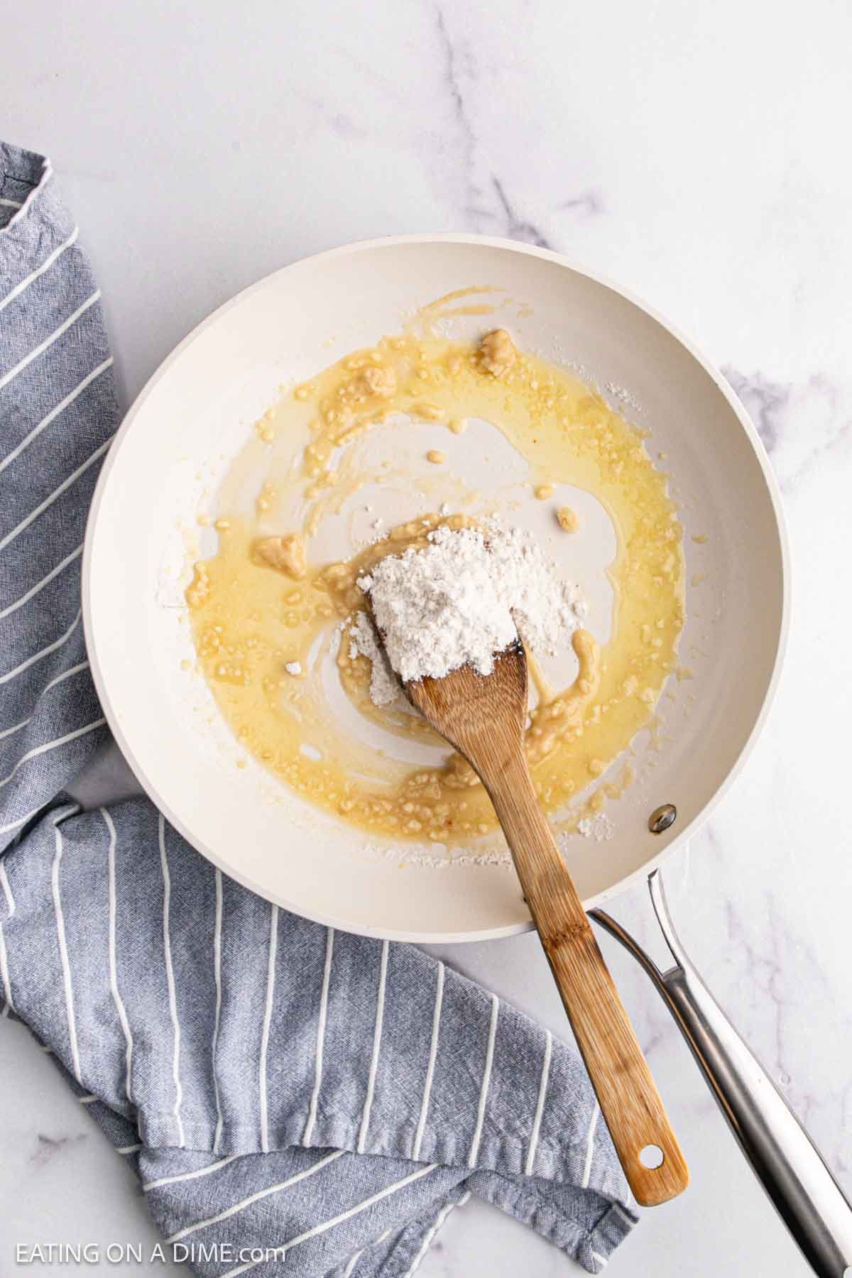 A white skillet contains melted butter and flour being mixed with a wooden spatula—the first step in making a creamy roux for Chicken Alfredo Bake. The pan rests on a marble surface next to a blue and white striped kitchen towel.