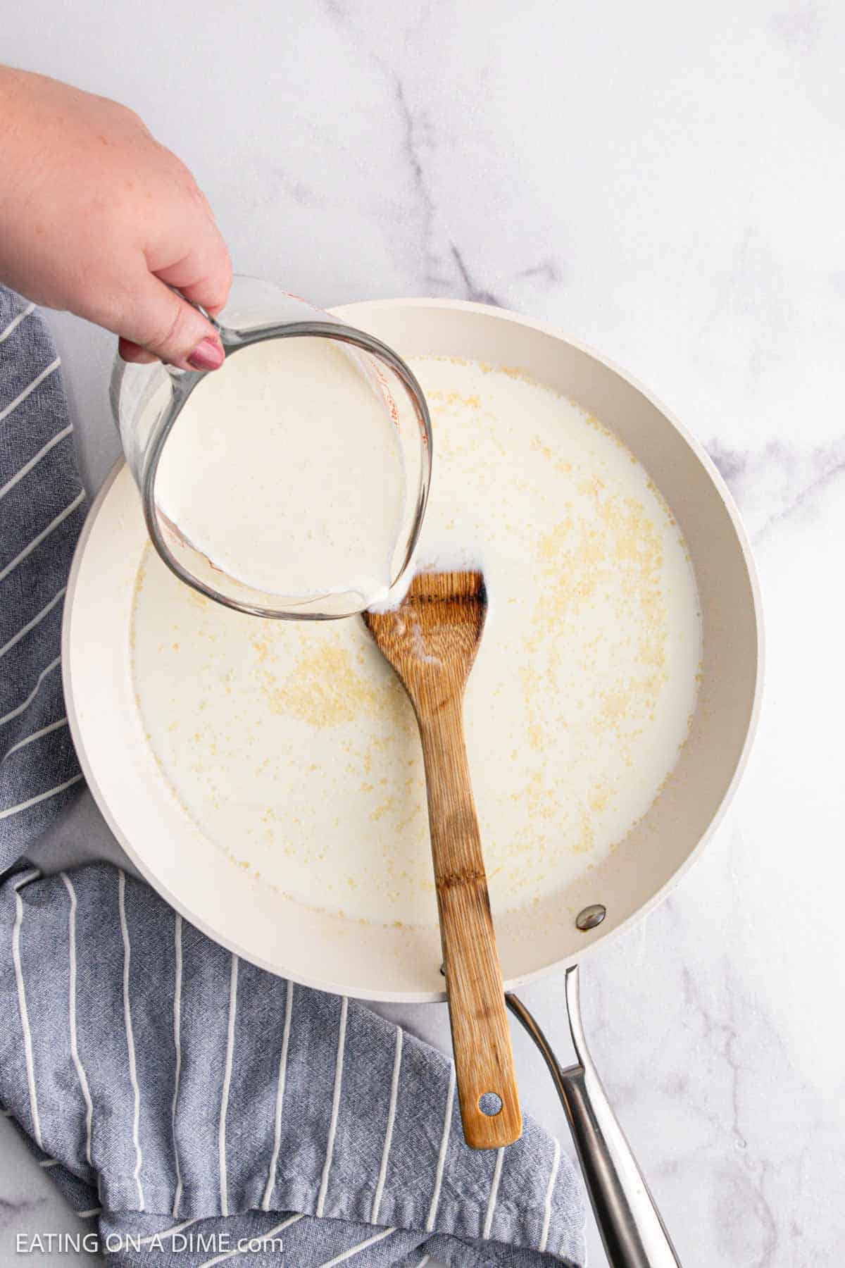 A hand pours cream from a glass measuring cup into a skillet with a wooden spatula inside, prepping the base for a delicious Chicken Alfredo Bake. The skillet rests on a marble surface next to a striped kitchen towel.
