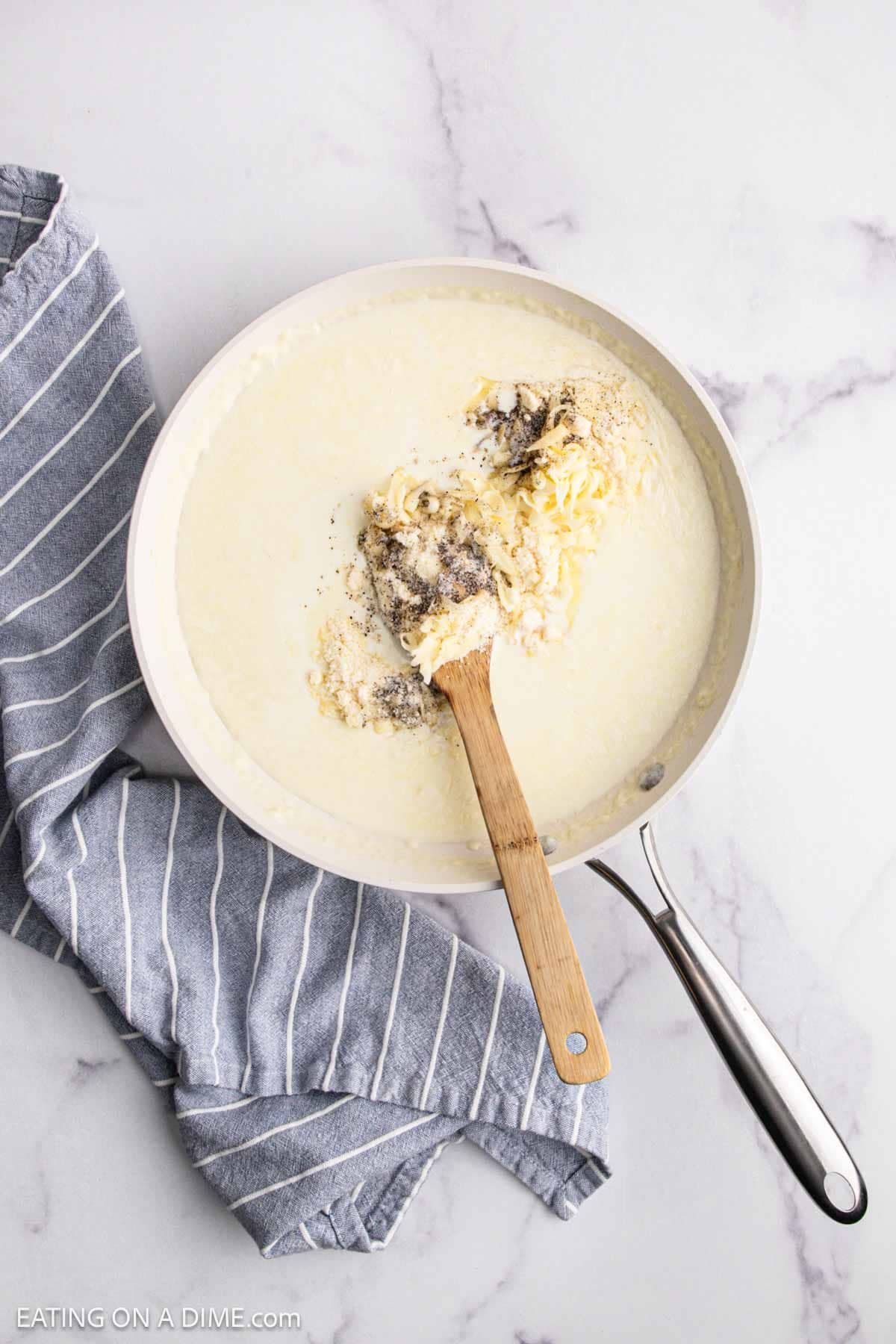 A saucepan filled with a creamy white sauce, shredded cheese, and black pepper—perfect for a Chicken Alfredo Bake—is being stirred with a wooden spoon. A blue and white striped kitchen towel rests beside the pan on a marble countertop.