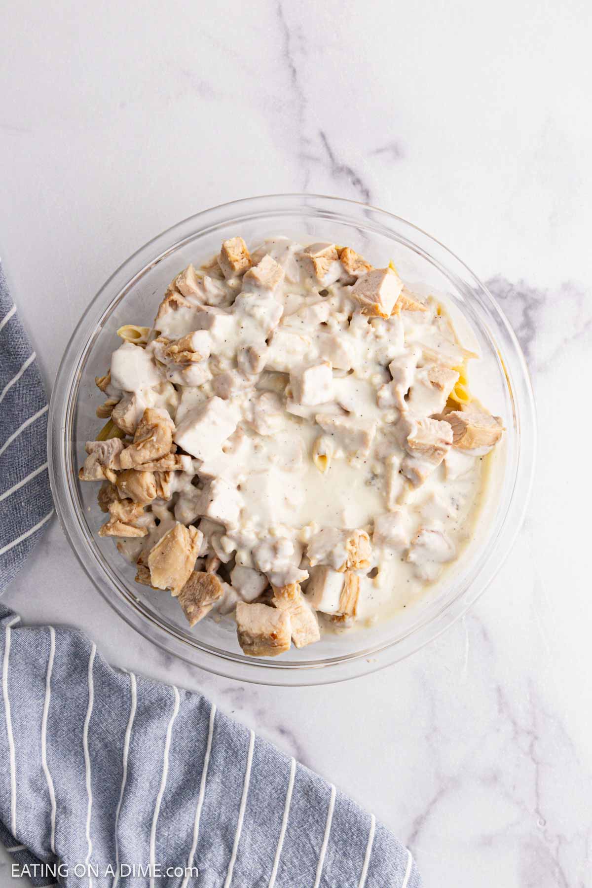 A glass bowl filled with creamy, diced Chicken Alfredo Bake sits on a white marble surface next to a blue and white striped kitchen towel.