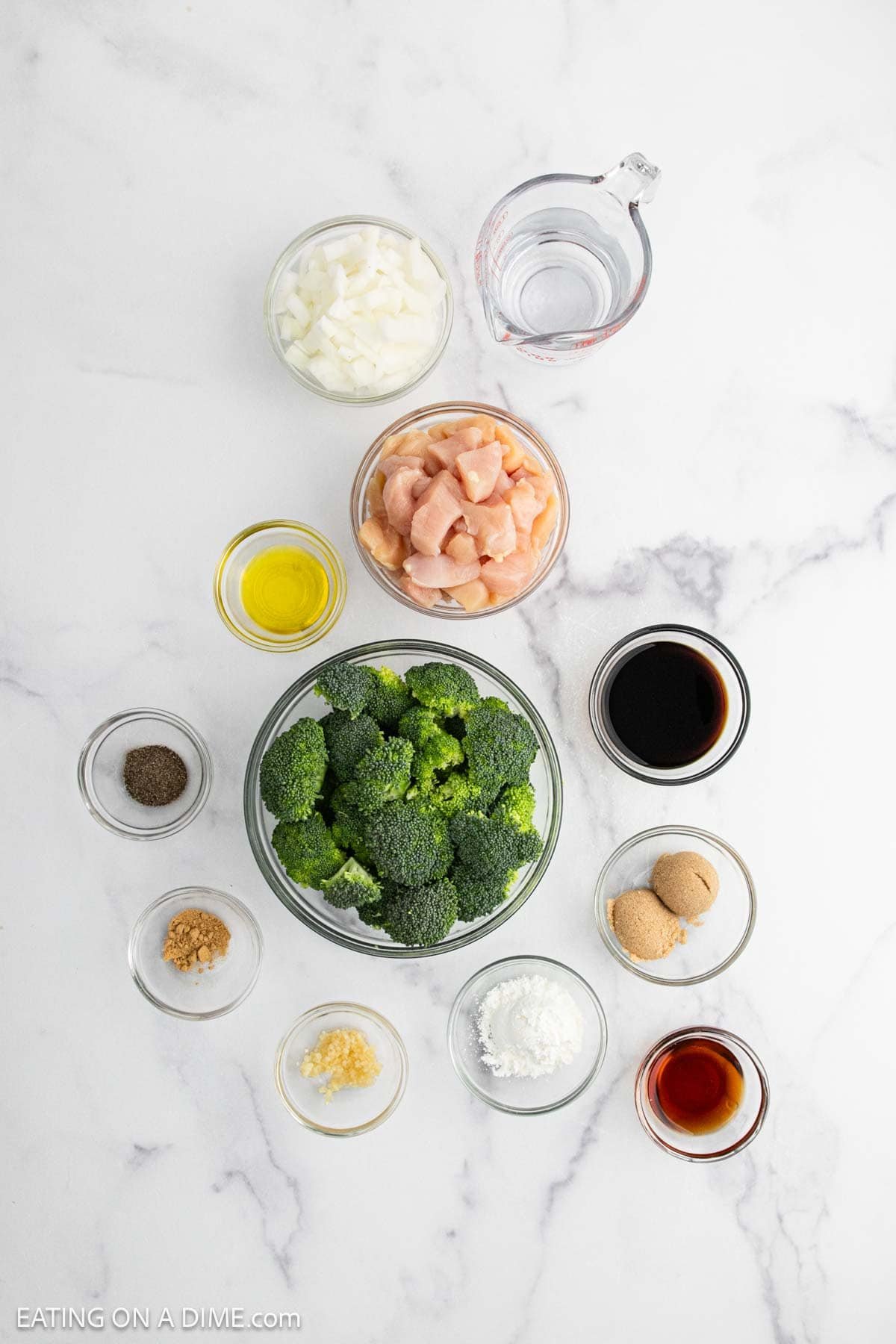 Overhead view of measured ingredients in small bowls on a white surface, ready to make a delicious chicken broccoli stir fry—featuring broccoli, diced chicken, chopped onions, soy sauce, brown sugar, olive oil, garlic, ginger, and more.