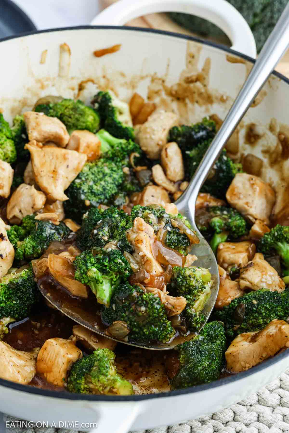 A close-up of a pan filled with chicken broccoli stir fry and onions in a savory sauce, with a large serving spoon scooping up a portion.