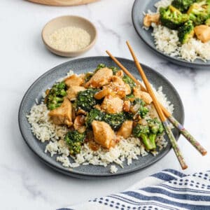 A plate of Chicken Broccoli Stir Fry sits atop white rice, garnished with sesame seeds and served with chopsticks. In the background, a dish of sesame seeds and another serving of this vibrant stir fry complete the scene.