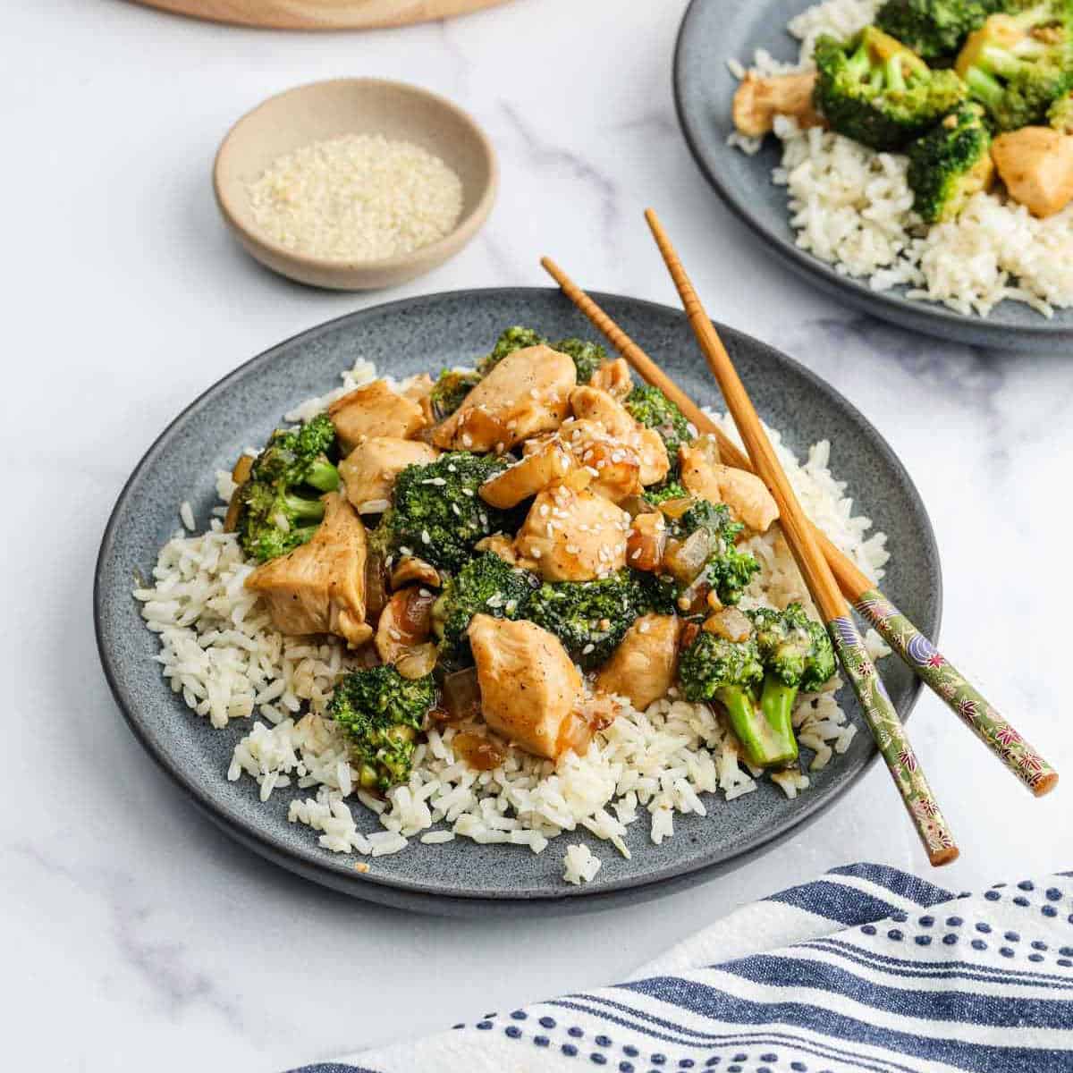 A plate of Chicken Broccoli Stir Fry sits atop white rice, garnished with sesame seeds and served with chopsticks. In the background, a dish of sesame seeds and another serving of this vibrant stir fry complete the scene.