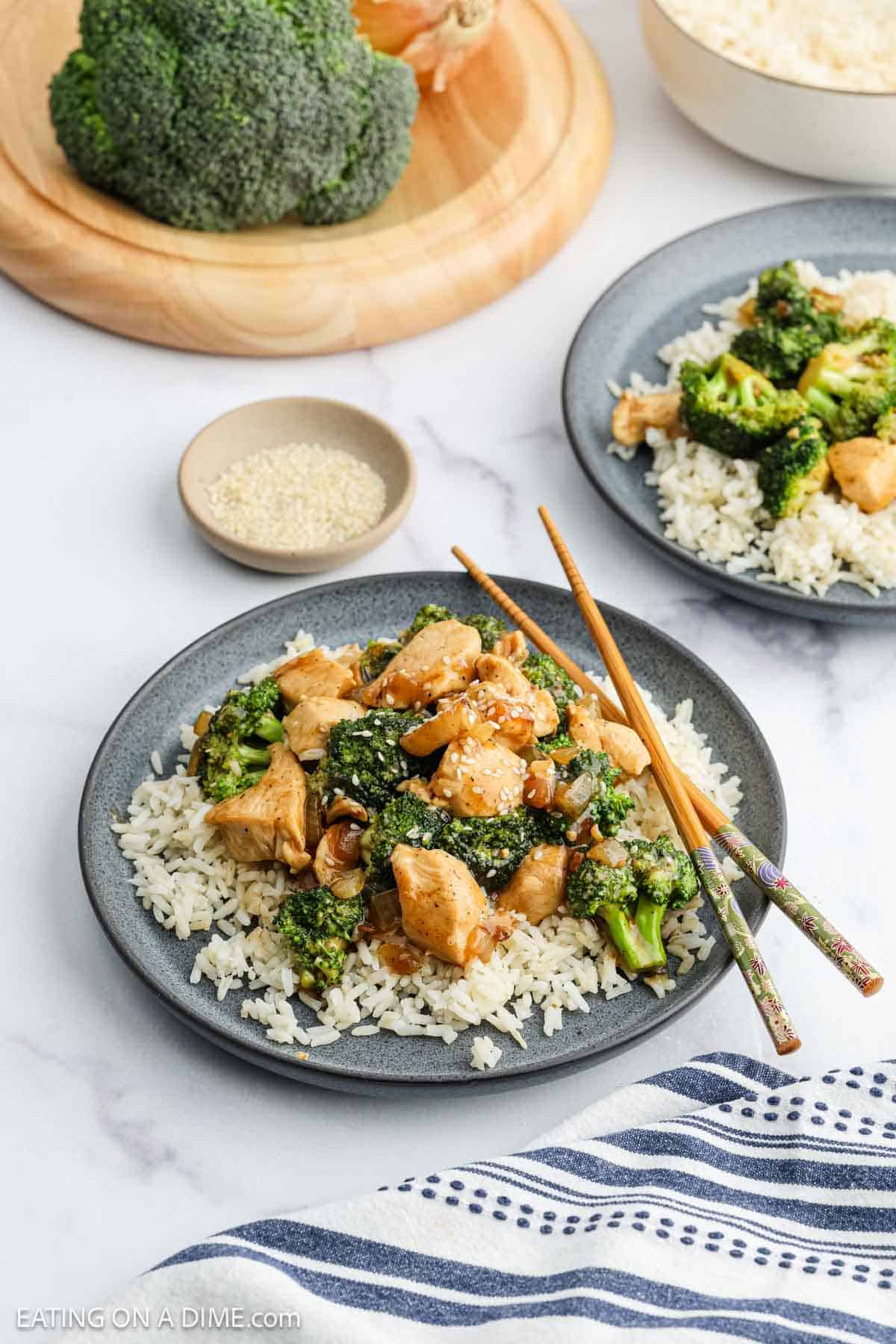 A plate of Chicken Broccoli Stir Fry sits atop white rice, garnished with sesame seeds. Chopsticks rest on the side, while extra broccoli, rice, and sesame seeds add to the inviting table spread.
