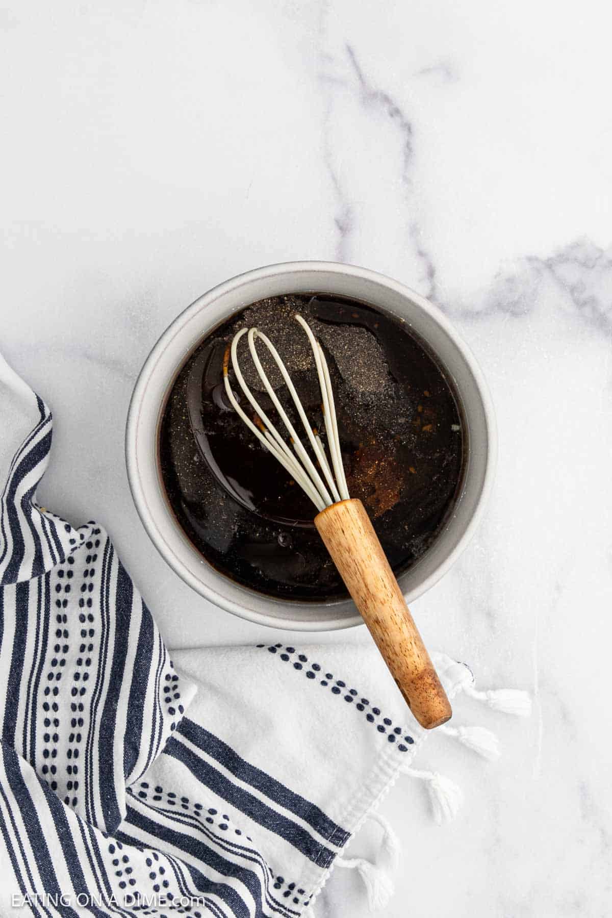 A bowl filled with a dark liquid marinade for chicken, topped with ground black pepper, and a small whisk with a wooden handle resting inside. A white and navy striped towel sits beside the bowl on a marble surface.