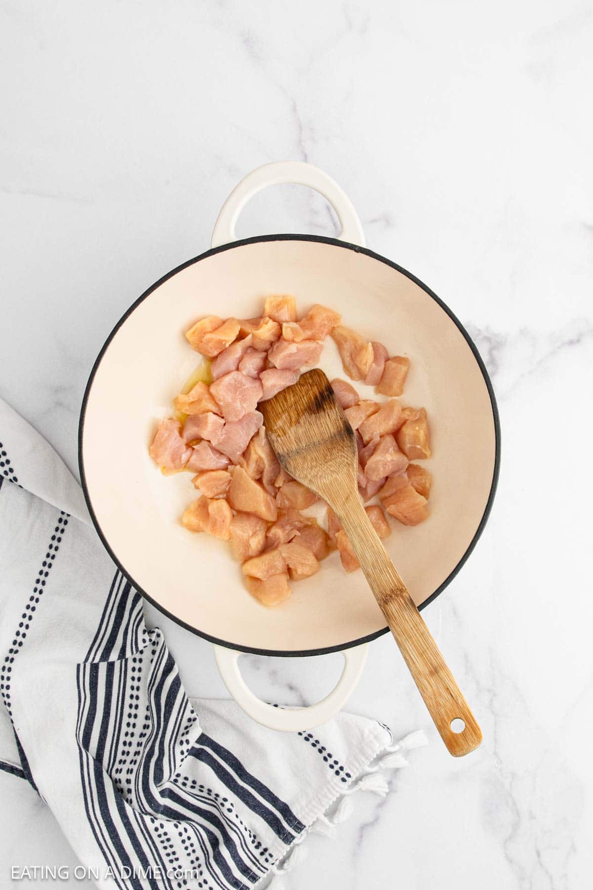 A white pot with raw, cubed chicken pieces for Chicken Broccoli Stir Fry and a wooden spatula inside sits on a marble surface next to a white and navy striped towel.