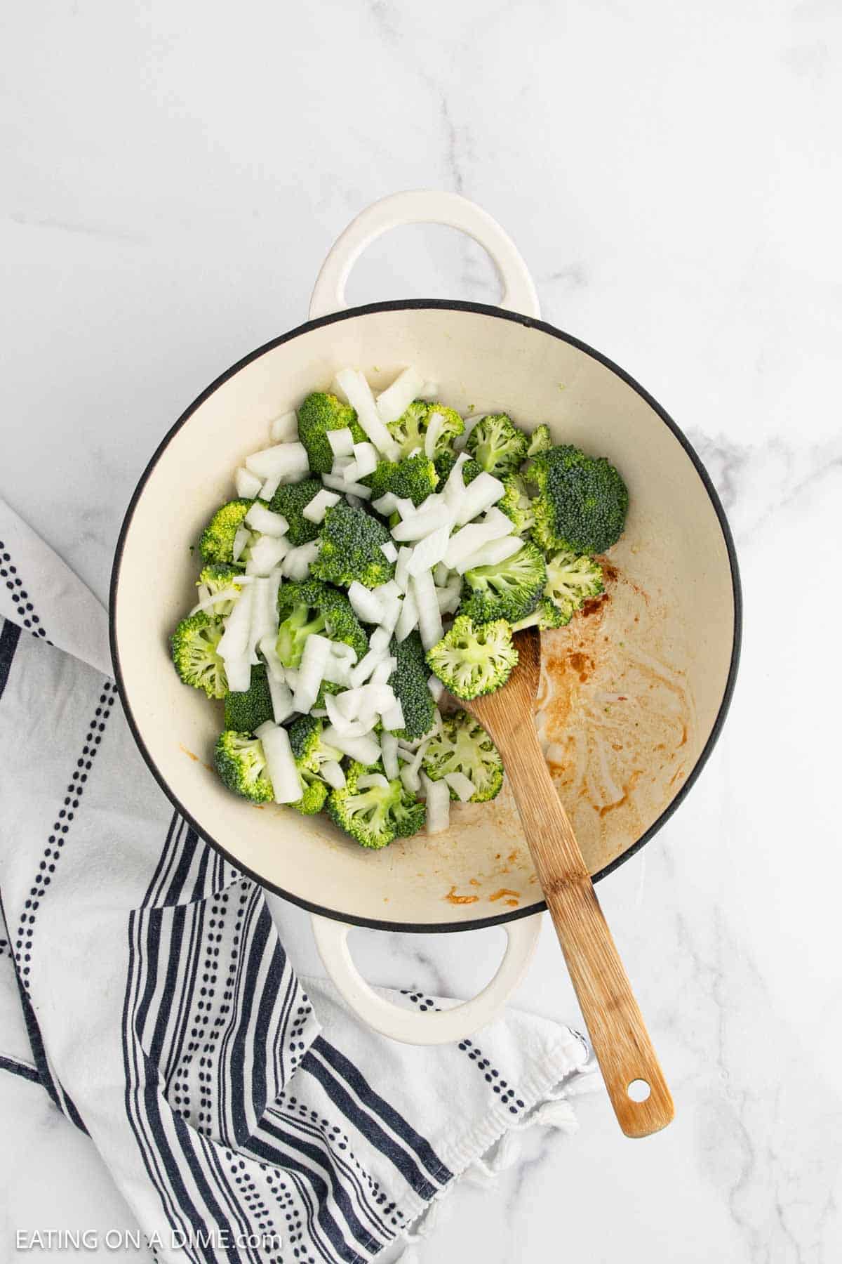 Overhead view of a white pot containing chopped broccoli and onions for a delicious Broccoli Stir Fry, with a wooden spoon. The pot sits on a white marble surface next to a black and white striped kitchen towel.