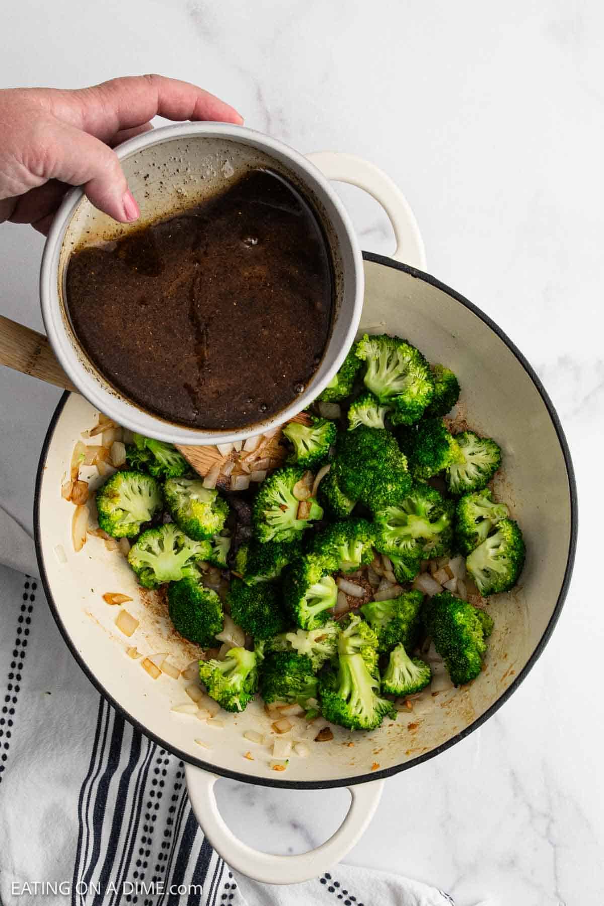 A hand pours a bowl of dark sauce into a pot filled with sautéed broccoli florets, chopped onions, and tender chicken on a white surface next to a striped towel—perfect for a flavorful stir fry.