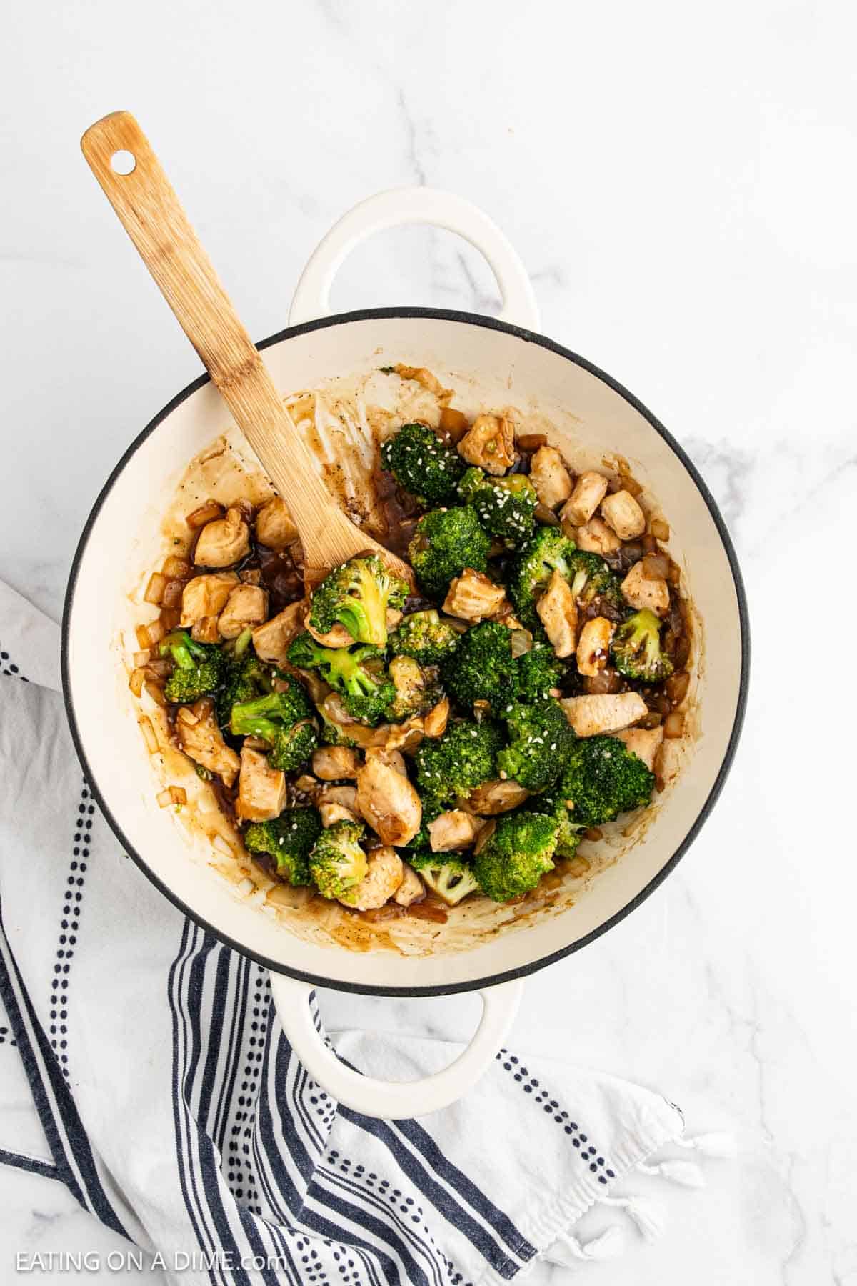 A white pot filled with Chicken Broccoli Stir Fry in a savory brown sauce, with a wooden spoon resting inside. The pot sits on a striped kitchen towel atop a white marble surface.