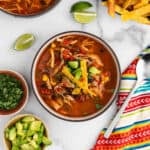 A bowl of Chicken Tortilla Soup topped with avocado chunks, crispy tortilla strips, and herbs sits surrounded by lime wedges, extra avocado, cilantro, and a colorful striped napkin atop a marble surface.