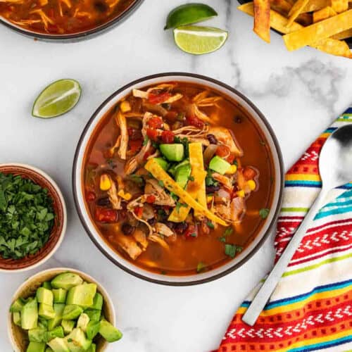 A bowl of Chicken Tortilla Soup topped with avocado chunks, crispy tortilla strips, and herbs sits surrounded by lime wedges, extra avocado, cilantro, and a colorful striped napkin atop a marble surface.