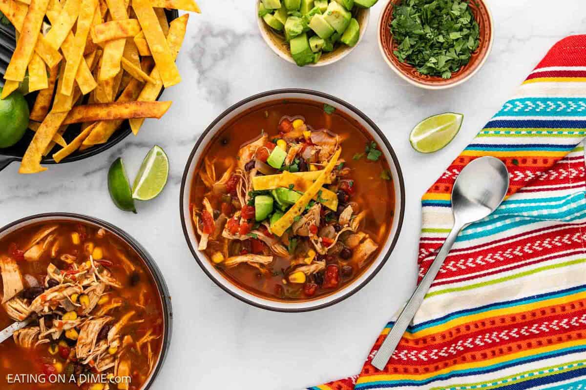 A bowl of Chicken Tortilla Soup topped with avocado, tortilla strips, and cilantro sits on a colorful striped cloth, surrounded by a spoon, extra tortilla strips, lime wedges, avocado, and cilantro—a perfect chicken soup recipe for any occasion.