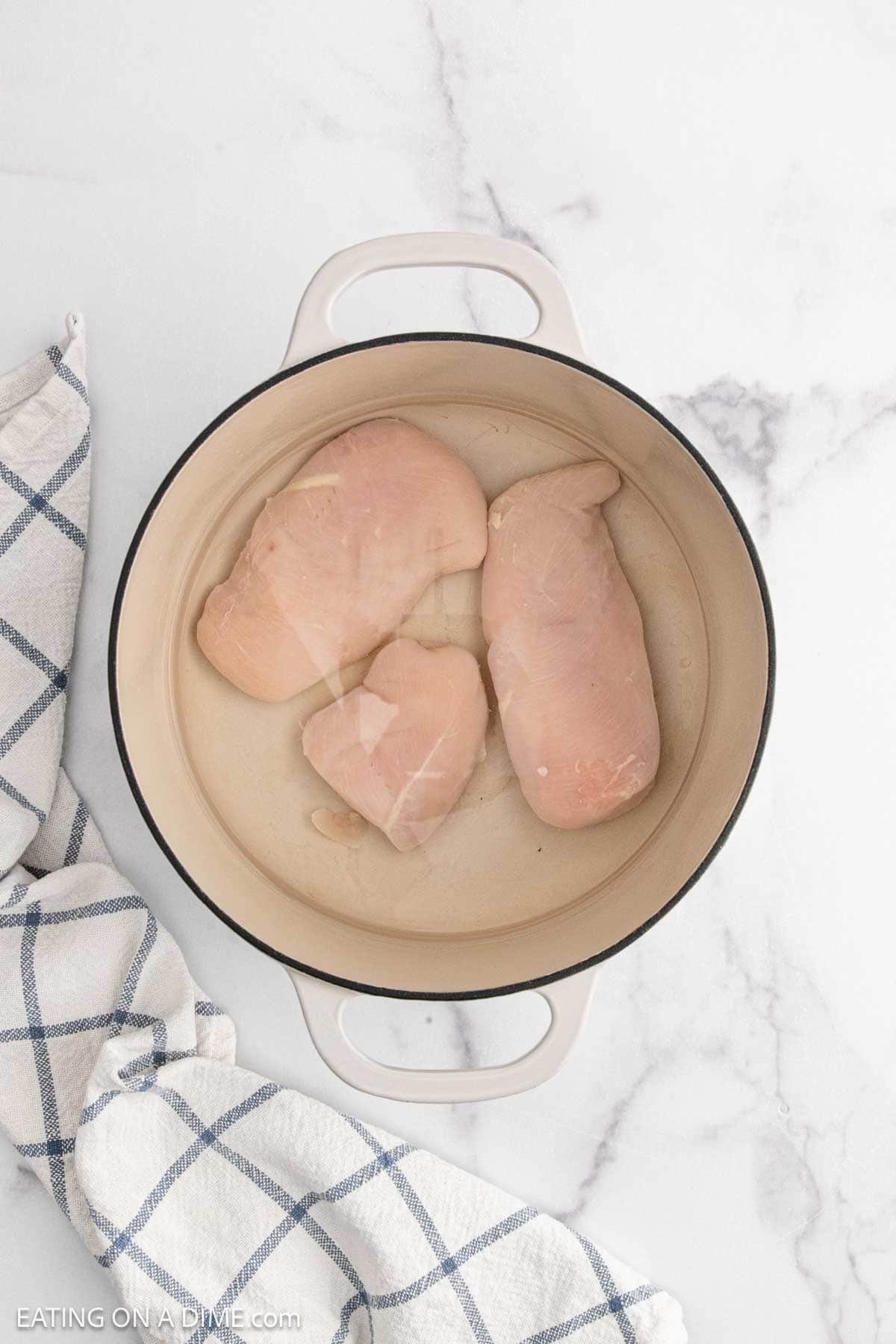 Three raw chicken breasts in a large beige pot of water, set on a white marble surface beside a blue and white checkered towel—perfect for starting homemade Chicken and Dumplings with Biscuits.