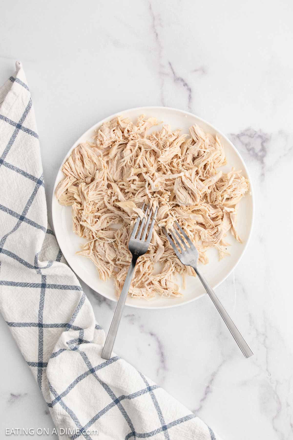 A white plate filled with shredded chicken, perfect for Chicken and Dumplings with Biscuits, two forks resting on the plate, and a white and blue checkered towel beside it on a marble surface.