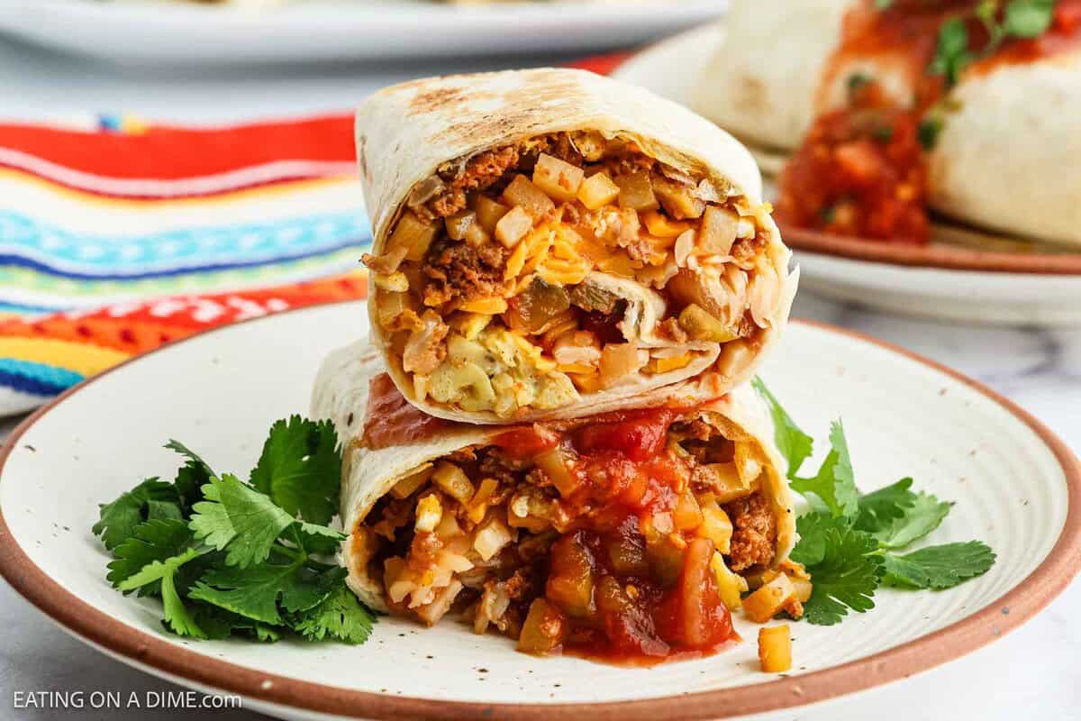 Two halves of a Chorizo Breakfast Burrito, filled with eggs, potatoes, cheese, and vegetables, are stacked on a plate with salsa and garnished with fresh cilantro. A colorful striped cloth is in the background.