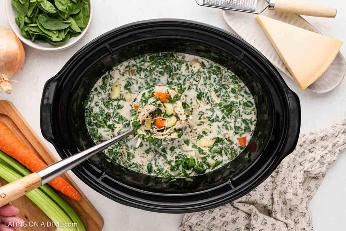 A Slow Cooker filled with creamy Chicken Gnocchi Soup, brimming with spinach, carrots, and herbs. Fresh spinach, celery, carrot, onion, and a block of cheese are arranged around the pot on a white countertop.