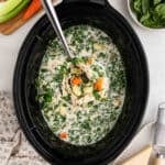 A Crock Pot brimming with creamy Chicken Gnocchi Soup, featuring shredded chicken, spinach, carrots, celery, and potatoes. A ladle lifts a hearty portion while fresh spinach and veggies rest nearby.