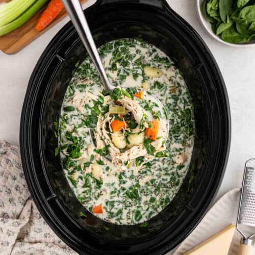 A Crock Pot brimming with creamy Chicken Gnocchi Soup, featuring shredded chicken, spinach, carrots, celery, and potatoes. A ladle lifts a hearty portion while fresh spinach and veggies rest nearby.