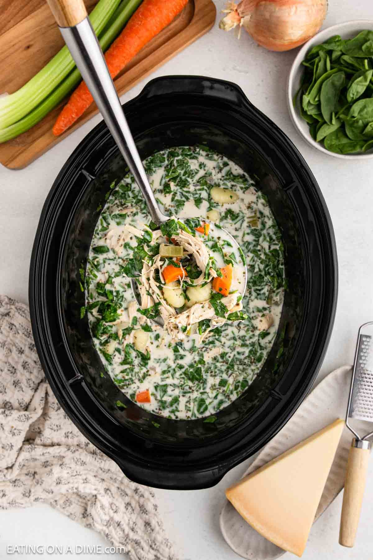 A ladle scoops creamy Chicken Gnocchi Soup from a black Crock Pot. Fresh carrots, celery, spinach, onion, and cheese are arranged nearby on a white countertop.