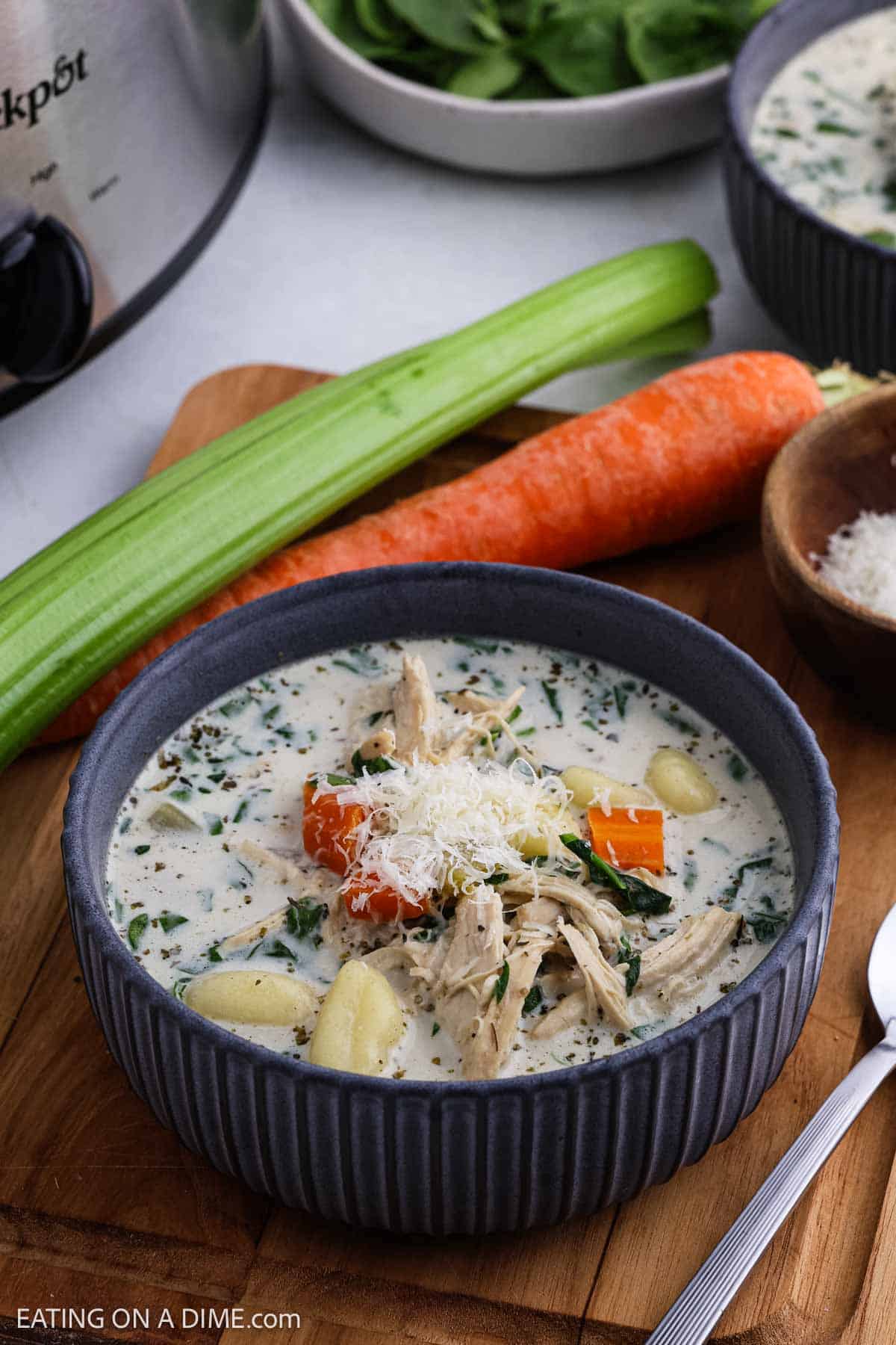 A bowl of creamy Crock Pot chicken and vegetable soup garnished with shredded cheese, featuring chunks of chicken, carrots, potatoes, and spinach. A carrot, celery stalk, and bowl of cheese appear in the background.
