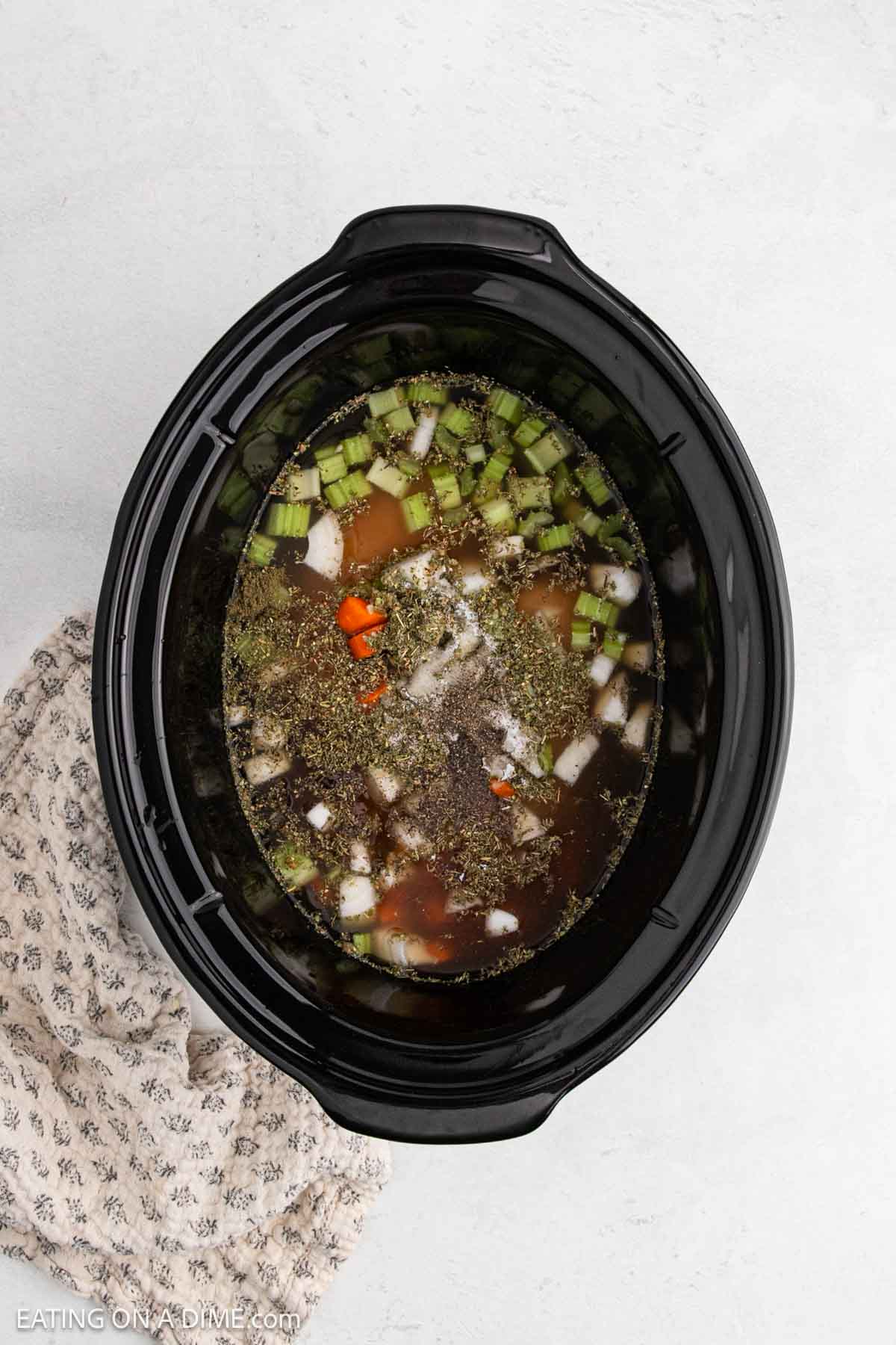 Overhead view of a Crock Pot filled with chopped vegetables, broth, and seasonings, ready to be cooked. A patterned kitchen towel is placed beside the slow cooker on a light surface. Perfect for making cozy Chicken Gnocchi Soup.