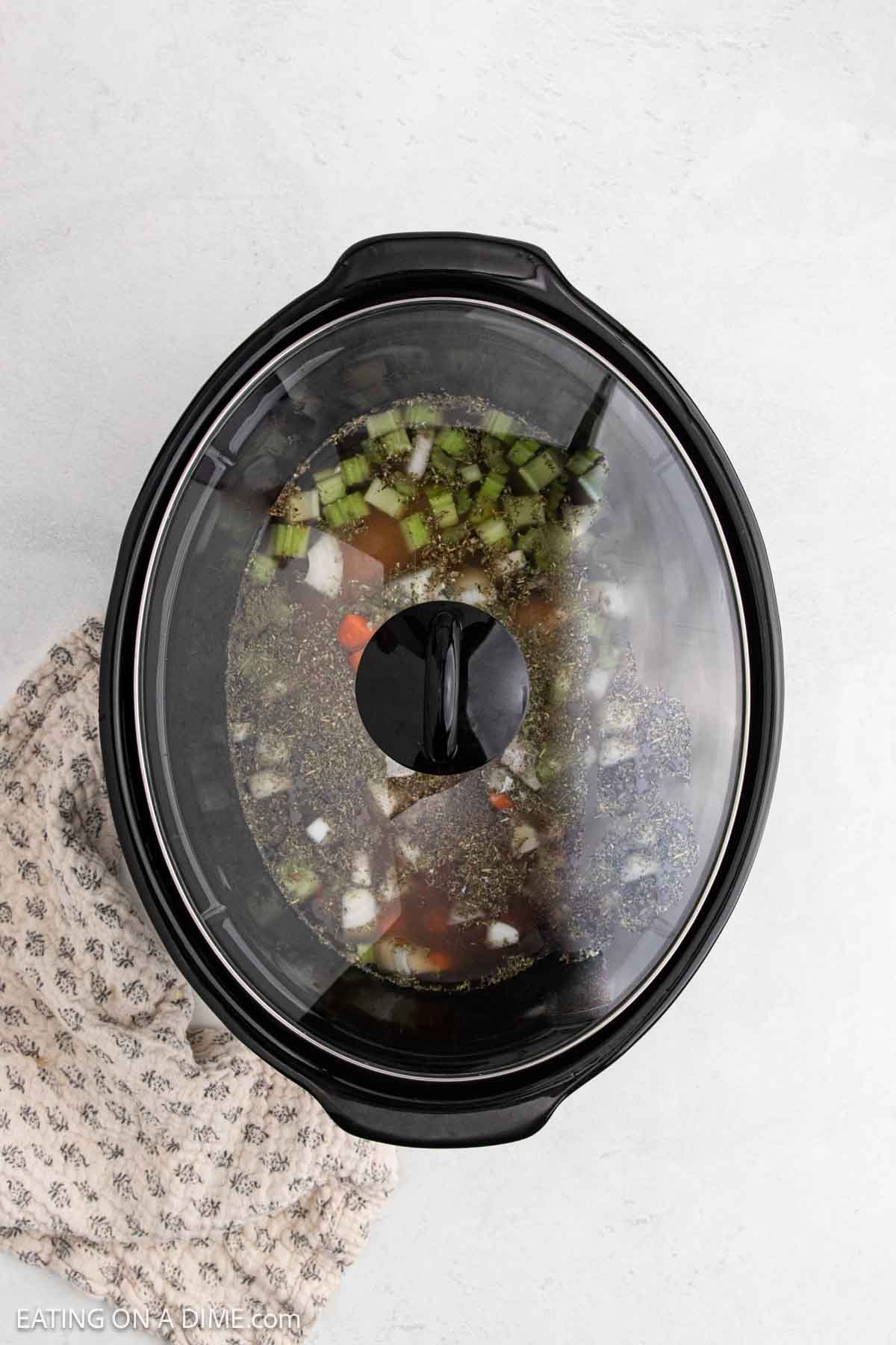 A Crock Pot filled with chopped onions, green peppers, tomatoes, and herbs simmers under a glass lid. Perfect for making comforting recipes like Chicken Gnocchi Soup, it sits on a light surface beside a patterned cloth.