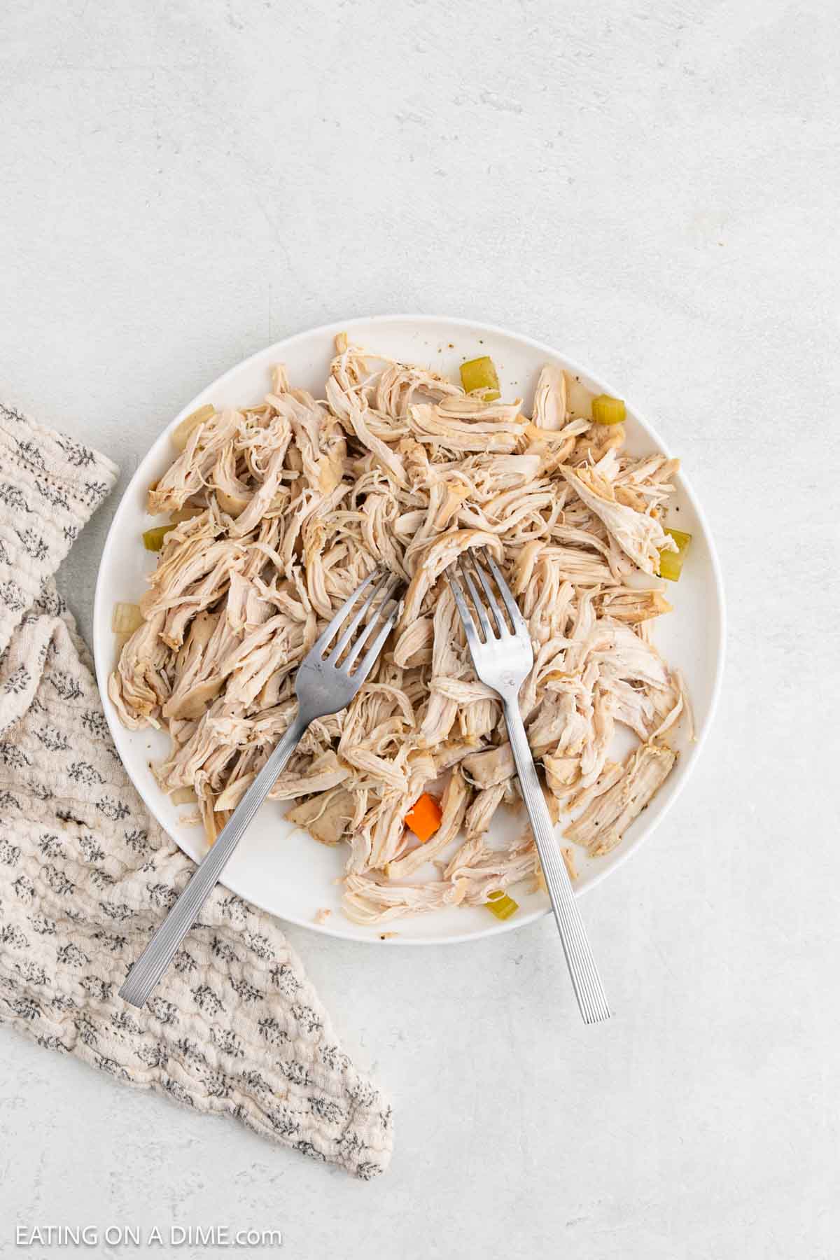 A white plate filled with shredded chicken and small vegetable pieces, reminiscent of Chicken Gnocchi Soup, with two forks placed on top. A textured beige towel is partially visible next to the plate.