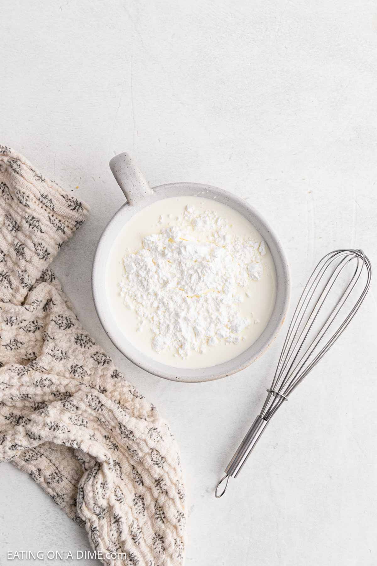A bowl filled with cream and powdered sugar sits next to a metal whisk and a textured beige cloth on a light surface, ready to become the perfect topping for your Slow Cooker desserts.