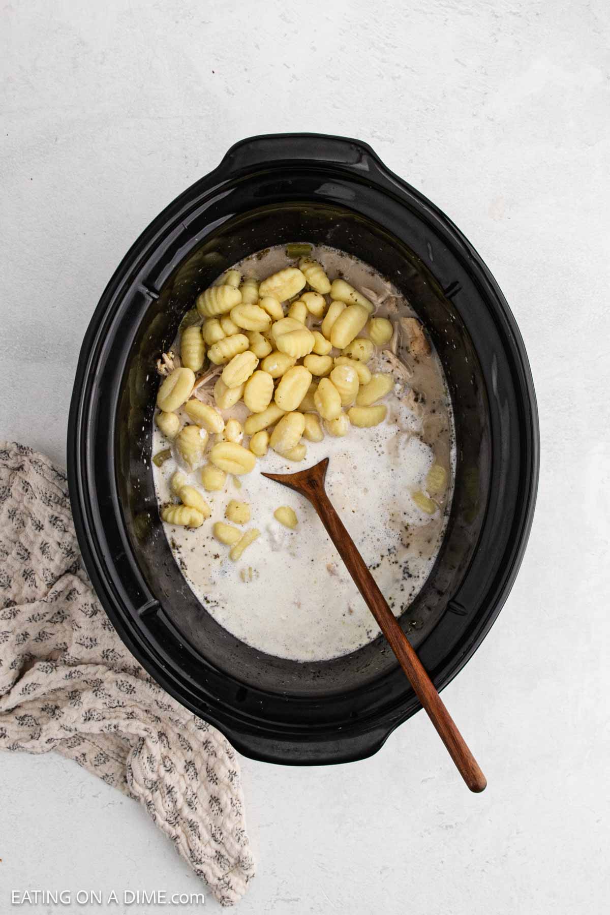 A black Crock Pot filled with creamy Chicken Gnocchi Soup and uncooked gnocchi, stirred with a wooden spoon. A textured beige kitchen towel rests next to the slow cooker on a light surface.